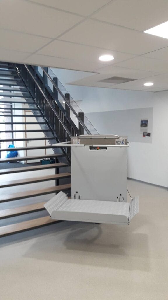 Plateautraplift met neergeklapt platform onderaan een stalen trap in een modern kantoorgebouw