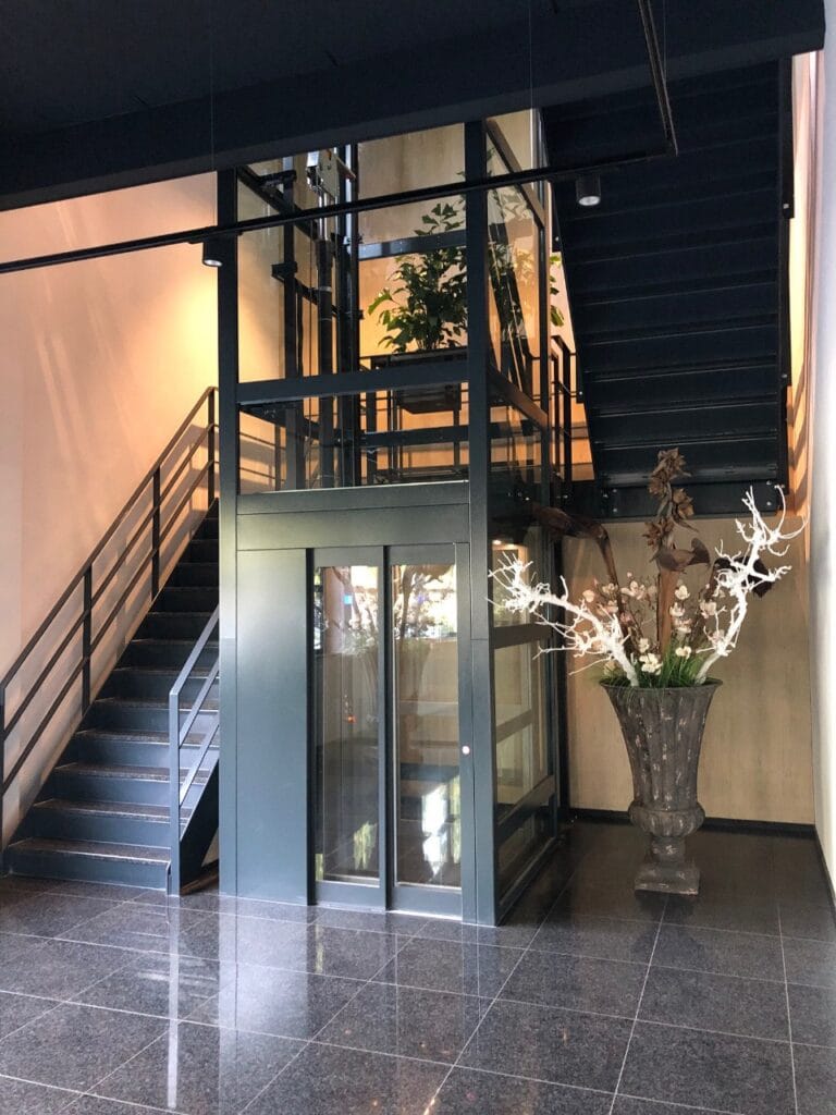 Glazen cabinelift met zwart stalen frame naast een trap in de lobby van Finder Relais Amsterdam