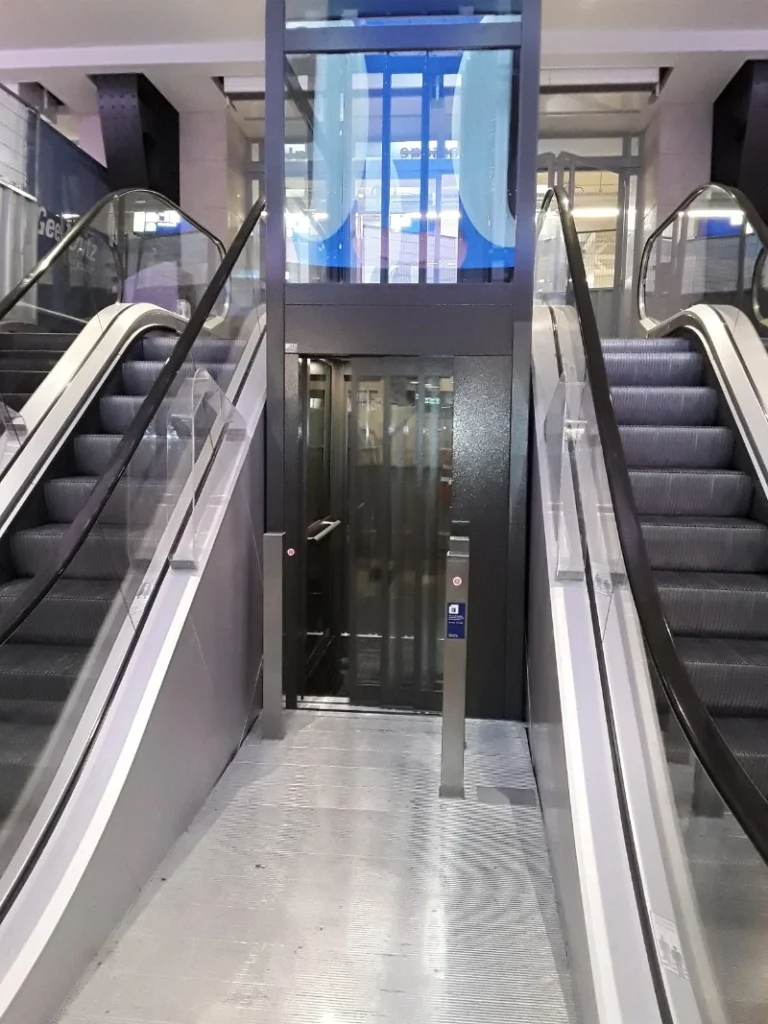 Glazen platformlift geplaatst tussen twee roltrappen in NS-station Amsterdam