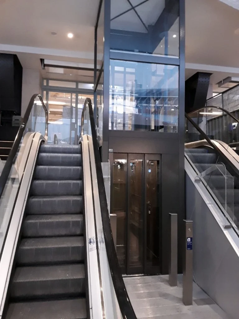 Glazen cabinelift met donker stalen frame tussen twee roltrappen in NS-station Amsterdam