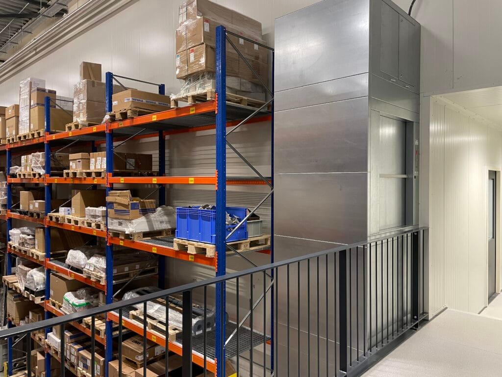 Goederenlift in roestvrijstalen uitvoering naast hoge stellingkasten in een magazijn