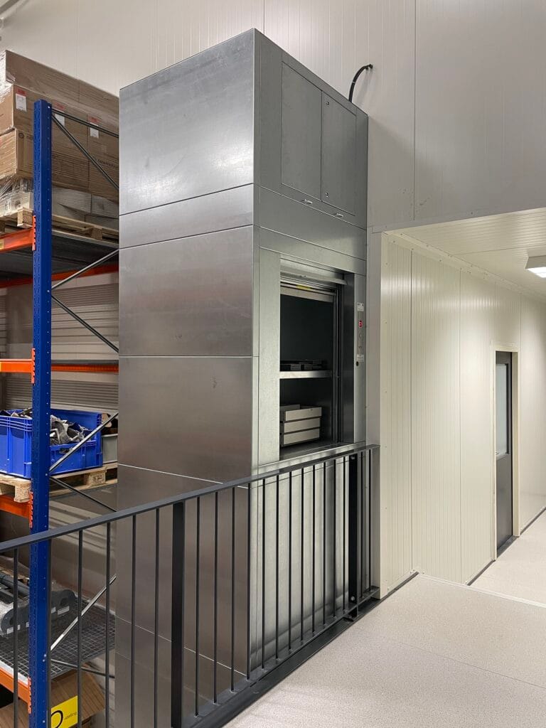 Goederenlift in roestvrijstalen behuizing op een magazijnverdieping met stellingkasten