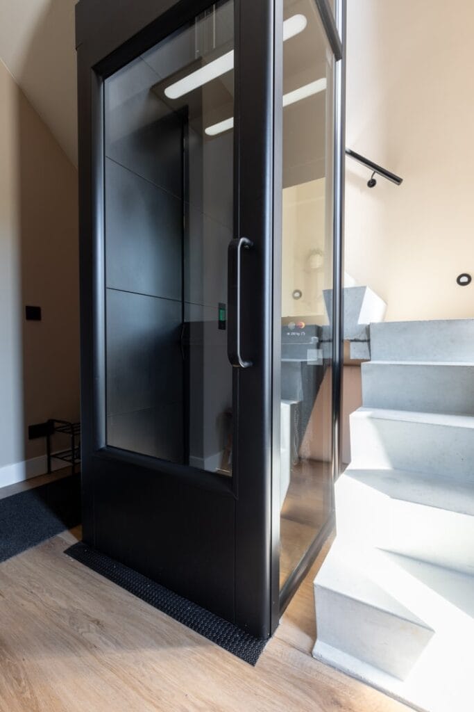Zwarte woonhuislift met glazen wanden naast betonnen trap in modern woonhuis