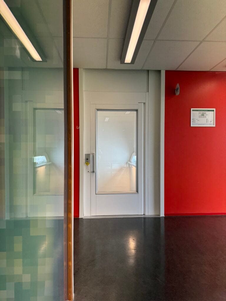 Witte platformlift met glazen deur in een gang met rode muur op het Spaarne College Haarlem