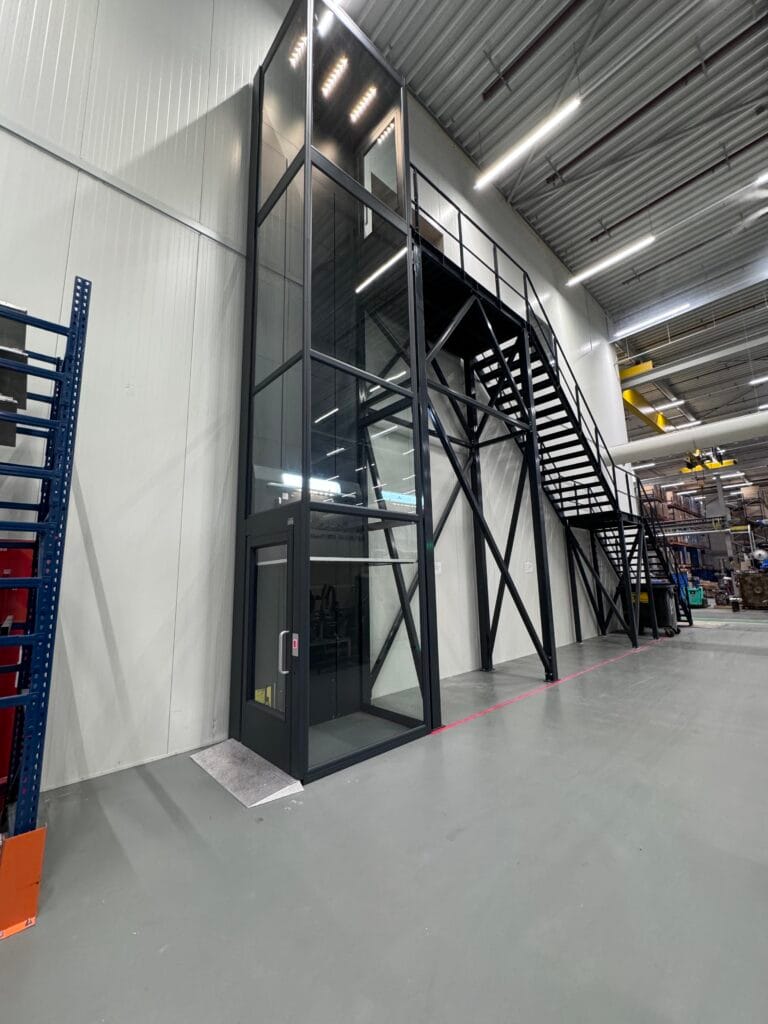 Glazen platformlift met zwart stalen frame naast een industriële trap in een magazijn