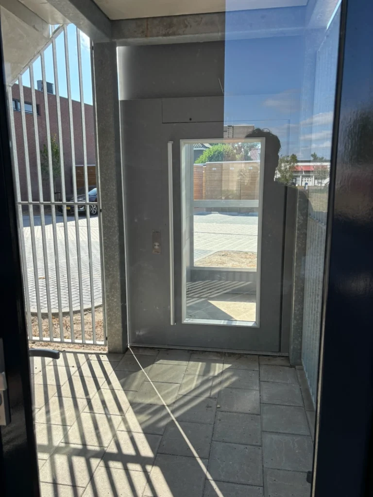 Platformlift met glazen deur geïnstalleerd in overdekte buitenruimte van appartementencomplex in Waalre