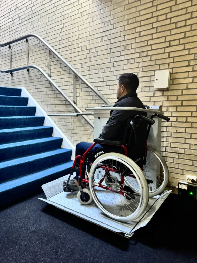 Rolstoelgebruiker op een plateautraplift naast een trap met blauwe vloerbedekking in een appartementencomplex in Roelofarendsveen