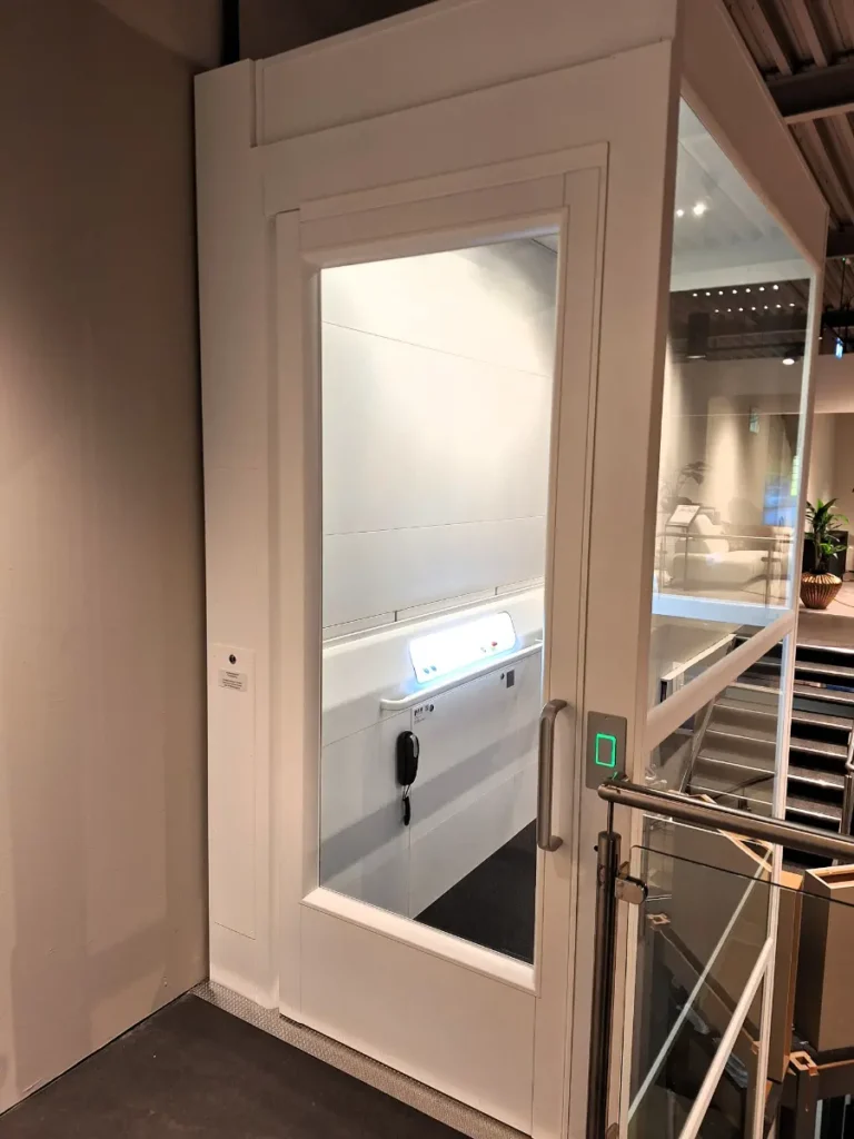 witte platformlift a 7000 met gesloten deur, gezien vanaf een vide naast een trap met glazen leuning in een woonwinkel