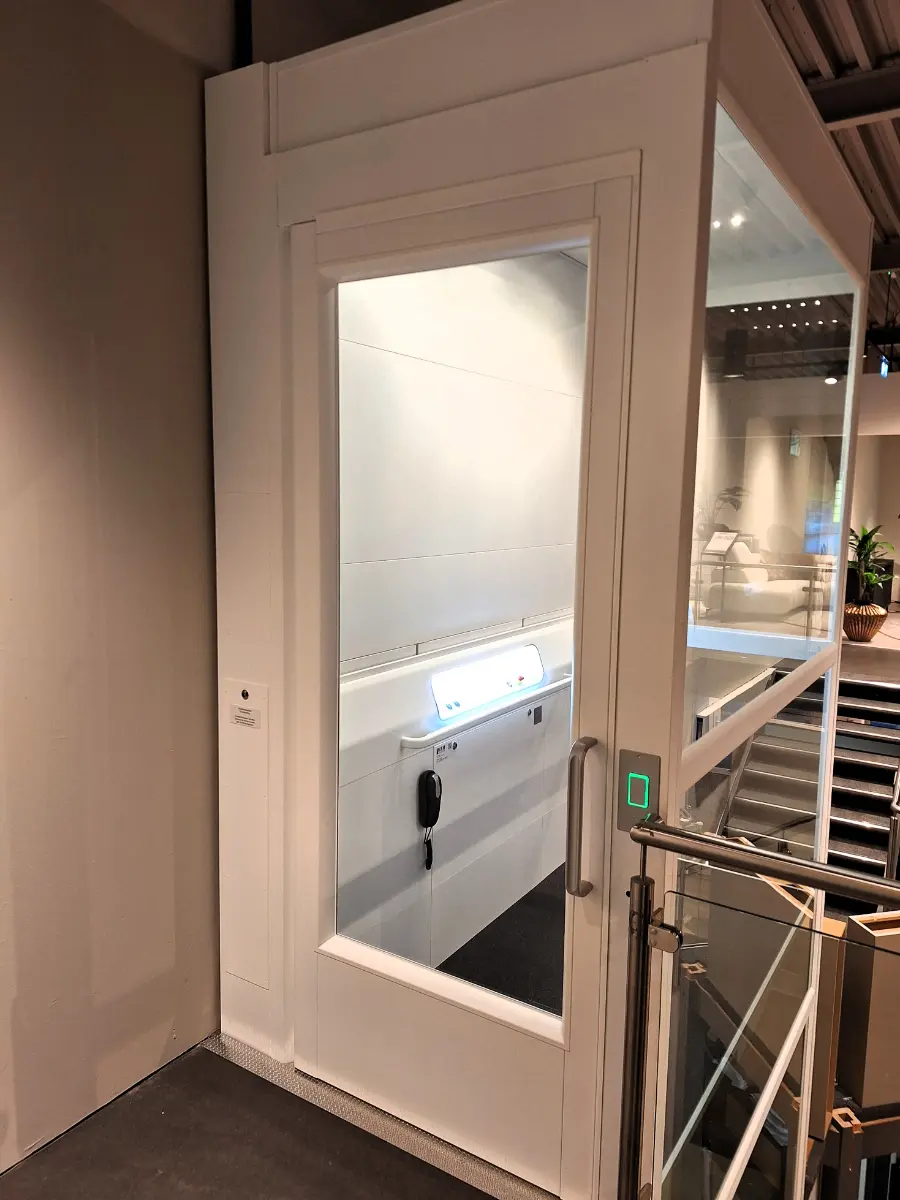 witte platformlift a 7000 met gesloten deur, gezien vanaf een vide naast een trap met glazen leuning in een woonwinkel